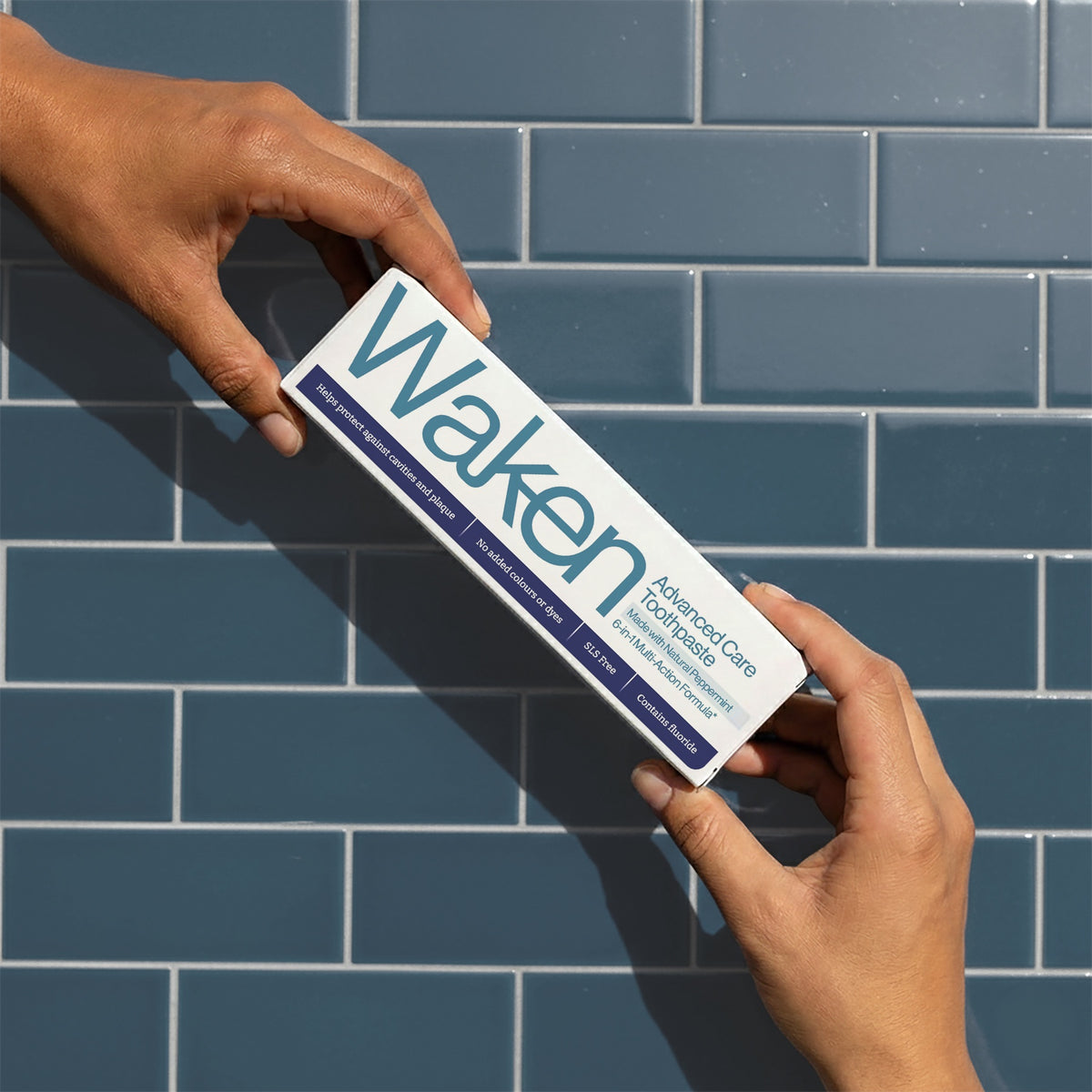 Hands passing a Waken Advanced Care toothpaste carton against blue tiles